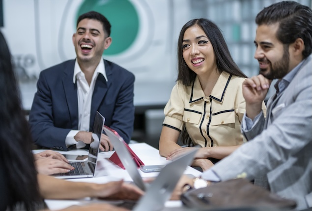 Employees sitting at a table with laptops, smiling at one another.